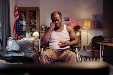 Siyahi bir adam televizyonun karşısında oturmuş sıkıcı TV programları izliyor ve elinde bir bardak alkol tutuyor.