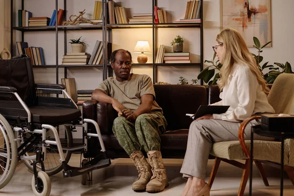 Black war veteran with disability sitting on couch and talking to psychologist listening to him