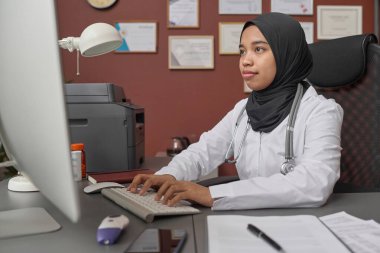 Hastane ofisinde oturan ve masaüstü bilgisayarında yazan tesettürlü genç bayan doktor.