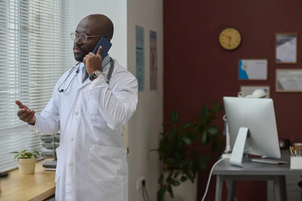African american doctor in lab coat talking on mobile phone standing at his workplace in office