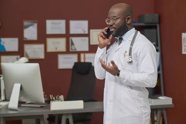 African american doctor in lab coat standing in office in hospital and having conversation on phone