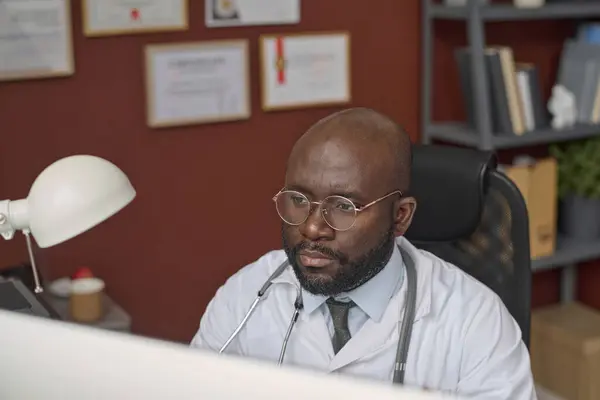 Black physician in lab coat and glasses attentively looking at computer monitor at his workplace