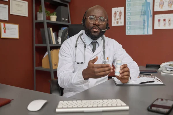 Black physician sitting at his workplace and showing pills bottle on webcam at virtual appointment