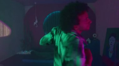 Genç enerjik dansçı kameraya bakıyor ve çok renkli neon ışıklarıyla karanlık stüdyoda hip hop hareketleri yapıyor.