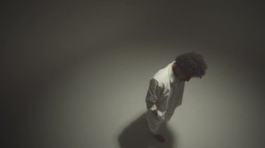 Yüksek açılı genç çıplak ayaklı, beyaz takım elbiseli, dans eden enerjik hip hop dansçısı stüdyoda spot ışıkları altında kameraya bakıyor.
