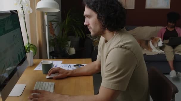 年轻的高加索商人在家里工作时 在台式计算机上分析商业图表和图表 他的伯明翰夫人和小猎犬坐在沙发上 背靠背 — 图库视频影像