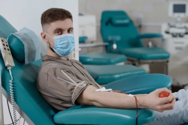 Koruyucu maskeli genç adam kan bağışı sırasında tıbbi sandalyede oturuyor ve kameraya bakıyor.