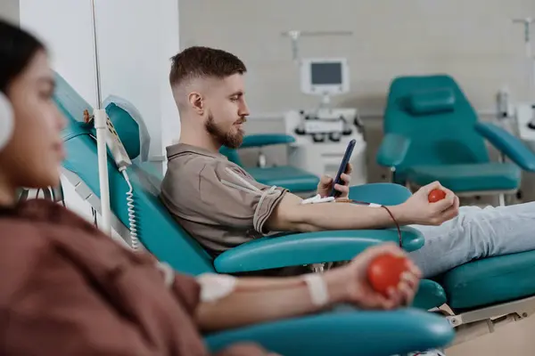 Caucasian man sitting in armchair during blood donation in hospital and scrolling his smartphone