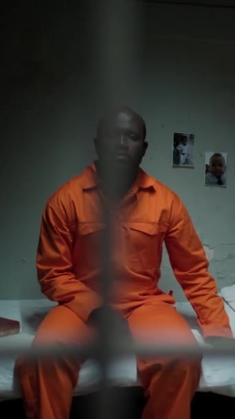 Pionowe Ujęcie Afroamerykanina Pomarańczowym Mundurze Siedzącego Łóżku Zamkniętą Ścianą Więziennej — Wideo stockowe