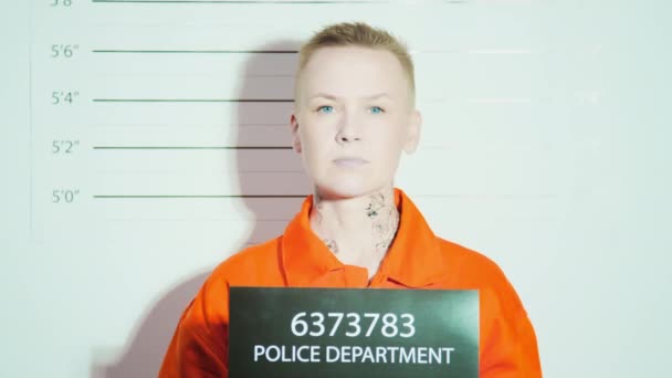 オレンジの刑務所の制服を身に着けているタトゥーを持つ短髪の女性は 刑務所のマグショットの高さチャートに対してカメラのためにサインを保持し ポーズします — ストック動画