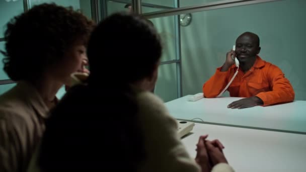 ガラスの壁の後ろに座っている黒人の囚人のラックフォーカスショットと刑務所で彼を訪問する家族との電話会話を持つ — ストック動画
