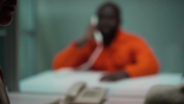 訪問室で会議中にガラスの壁の後ろに座っている投獄された夫と電話で話す母親と子供のショットの肩のラックの焦点の上に — ストック動画