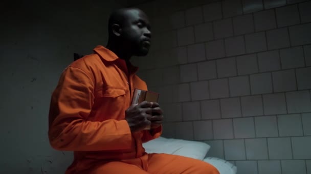 African American Inmate Sitting Bed Prison Cell Holding Bible Close — Stock Video
