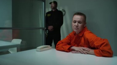 Turuncu hapishane üniformalı kadın mahkum hapishane ziyaretçi odasında oturuyor ve üzgün bir şekilde kameraya poz veriyor, arka planda nöbet tutuyor.