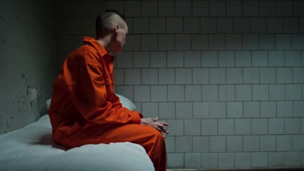 壁に割れたペンキで暗い刑務所のセルでベッドに座っているオレンジ色の制服の女性のコピースペースで中長いショット — ストック動画