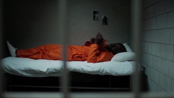 穿着橙色制服的非洲裔美国囚犯躺在牢房的床上 看着墙上的家人照片 却错过了他们 纵观他们的酒吧 — 图库视频影像