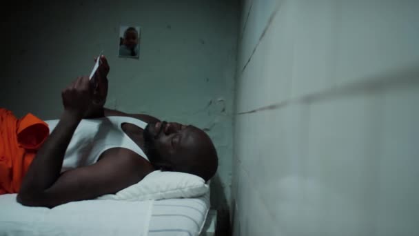 Несчастный Чернокожий Заключенный Лежит Кровати Тюрьме Смотрит Фото Ребенка Скучает — стоковое видео