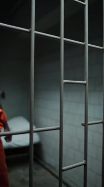 Afro-Amerikalı bir mahkumun hapishane hücresinde parmaklıkları tutarken ve kameraya bakarken dikey görüntüsü.