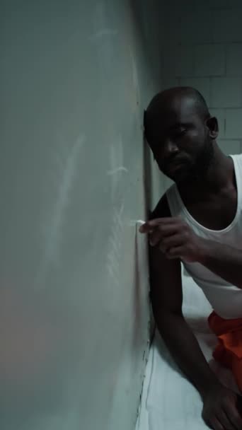 在牢房里躺在床上的悲伤黑人囚犯被垂直拍摄 并在墙上画上记分牌 同时数着释放前的日子 — 图库视频影像