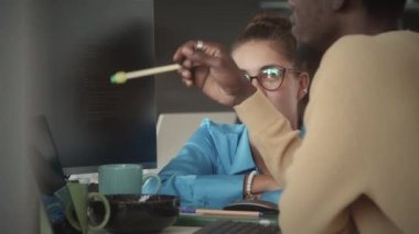 Genç beyaz kadın yönetici, ofiste işbirliği yaparken Afrika kökenli Amerikalı bilişim uzmanıyla bilgisayar ekranında programlama kodunu tartışıyor.