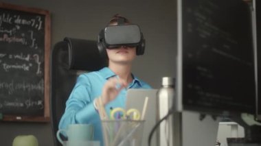 VR kulaklıklı genç bayan programcı, ofiste yeni uygulamayı test ederken dizüstü bilgisayarla oturup görünmez dokunmatik ekrana dokunuyor.