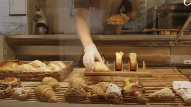使い捨て可能な手袋で認識できないパン屋のクロップショットは 販売のための甘いペストリーとガラスディスプレイケースの中でおいしいクロワッサントを置きます — ストック動画