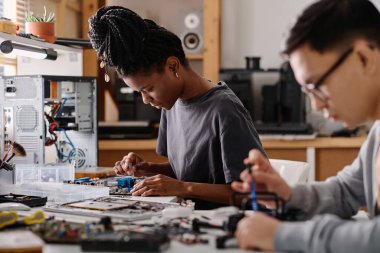 Siyah kadın tamirci, elektronik cihazı tornavidayla tamir ediyor. Erkek arkadaşı da yakınlarda çalışıyor.