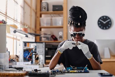 Dişi Afrikalı Amerikalı teknisyen dürbünlü gözlük takıyor ve işlemciyi dikkatle inceliyor.