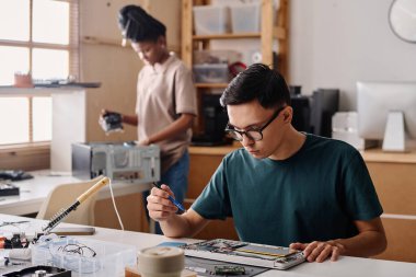 İş için kablosuz lehim kullanan genç teknisyen, arka planda duran siyah meslektaşı