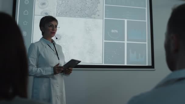 穿着白衣的女医生在主持医学会议时使用数码平板电脑 并在听众面前用麦克风说话 — 图库视频影像