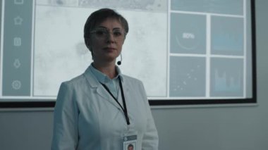 Tıbbi konferans sırasında mikroskop görüntüleri ve grafiklerle projeksiyon ekranının önünde kameraya poz veren beyaz önlüklü kadın doktor portresi