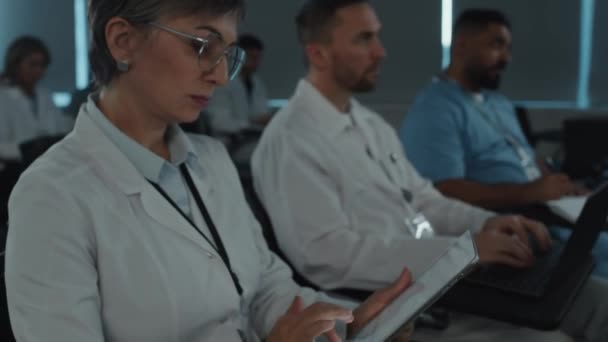 医疗会议期间 穿着白衣的女医生仔细听演讲人讲话和在数码平板电脑上打字时的倒立镜头 — 图库视频影像