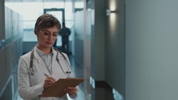 穿着白衣的中年女医生的画像 在剪贴板上写下笔记 并在医院走廊摆好姿势拍照 — 图库视频影像