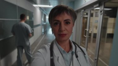 Beyaz önlüklü, kablosuz kulaklıklı, hastane koridorunda yürüyen ve video ararken ya da video kaydederken kameraya konuşan olgun kadın doktorların POV 'si.
