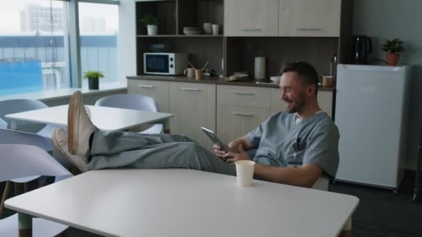 テーブルの上に足で座って デジタルタブレットでソーシャルメディアビデオを見て 休憩室でコーヒーを飲んでリラックスした医療ユニフォームの医療従事者 — ストック動画