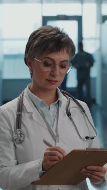 穿着白衣 脖子上戴听诊器 在剪贴板上写下便条 在医院工作时看着相机的专业女医生的画像 — 图库视频影像
