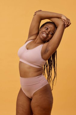 Kameraya poz veren rahat iç çamaşırı giyen neşeli Afrikalı Amerikalı kadının orta boy stüdyo resmi, sıcak kayısı rengi arka plan.