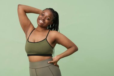 Rahat giyimli, gülümseyen ve kameraya poz veren, kendine güvenen siyahi bir kızın orta ölçekli stüdyo portresi.