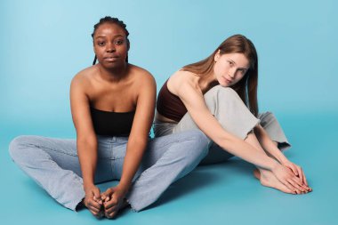 Etnik açıdan farklı iki genç kadın gündelik giysiler giyip stüdyoda rahatça oturup gökyüzü mavisi arka planda kameraya bakıyorlar.