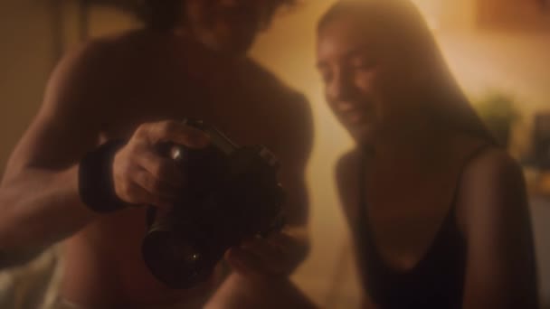 ガールフレンドにデジタルカメラに写真を表示し 寝室でロマンチックな家の写真撮影の後 彼女と話し合う若いシャツレスの男 — ストック動画