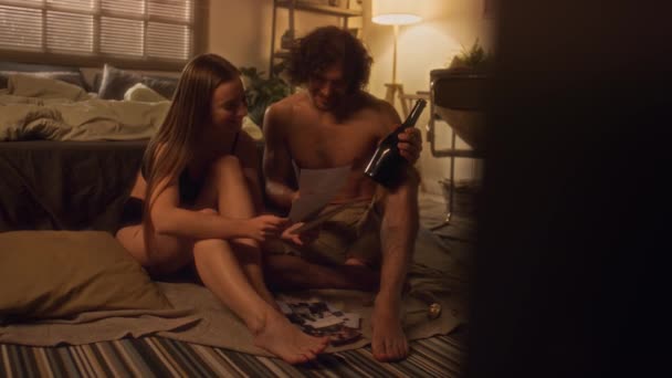 若いカップルは 暖かい光で居心地の良い寝室で床に座って 写真を見て ワインを飲んで 自宅でロマンチックなデートの夜を過ごしながらチャット — ストック動画