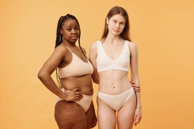 Kameraya poz veren iki özgüvenli Z-Siyah ve Kafkasyalı kadının orta uzunlukta stüdyo portresi.