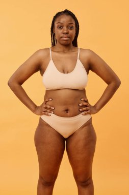Minimalist iç çamaşırı giyen, elleri kalçasında kameraya poz veren kendine güvenen siyahi bir kadının orta boy stüdyo portresi.
