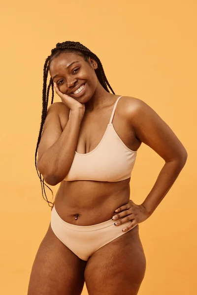 Studio portrait of modern young African American woman with braided hair wearing comfortable lingerie posing for camera, warm apricot background
