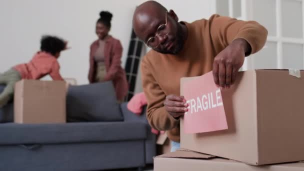 アフリカ系アメリカ人男性は ボール紙の箱に脆弱な看板を貼り バックグラウンドで遊ぶための荷物を詰めている間 — ストック動画