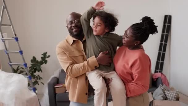 アフリカ系アメリカ人の両親が 小さな息子を腕に抱き 移動後に荷物を詰めずに新しい家で笑顔でカメラのために一緒にポーズする喜びの肖像 — ストック動画