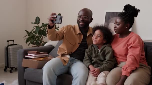 陽気なアフリカ系アメリカ人の家族がリビングルームでソファーに座り 新しいアパートで移動中に携帯電話でビデオ通話でチャット — ストック動画