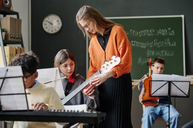 Genç beyaz bir kadının müzik dersinde genç bir kıza bas gitar çalmayı öğrettiği orta boy bir fotoğraf.