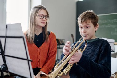 Müzik dersinde trompet çalan beyaz bir öğrencinin orta boy fotoğrafı. Bayan öğretmeni onu dinliyor.