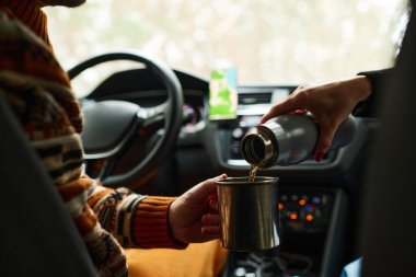 Tanımlanamayan bir kadın arabada oturmuş kocası için elektrikli süpürgeden bardağa sıcak çay dolduruyor.